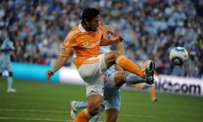 Brian Ching, con la camiseta naranja del Houston Dynamo, lanza una patada con la pierna derecha para controlar el balón ante la marca de un rival.