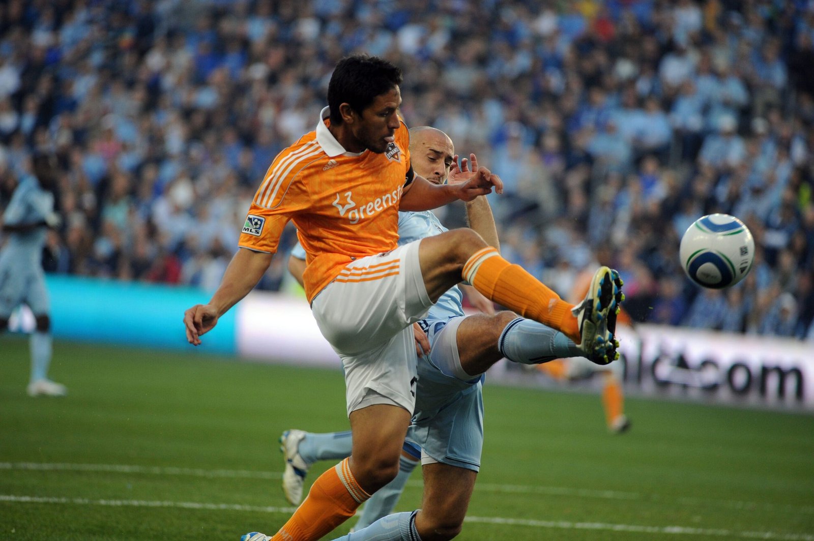 Brian Ching, con la camiseta naranja del Houston Dynamo, lanza una patada con la pierna derecha para controlar el balón ante la marca de un rival.
