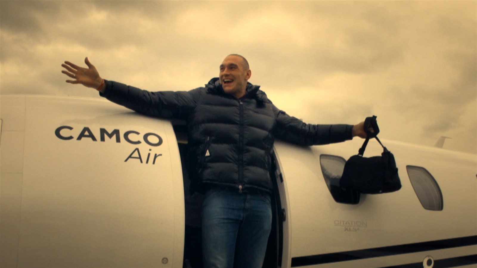 Escena del tráiler del documental TYSON FURY, de Netflix: un hombre sonrÃe con los brazos abiertos junto a un avión privado