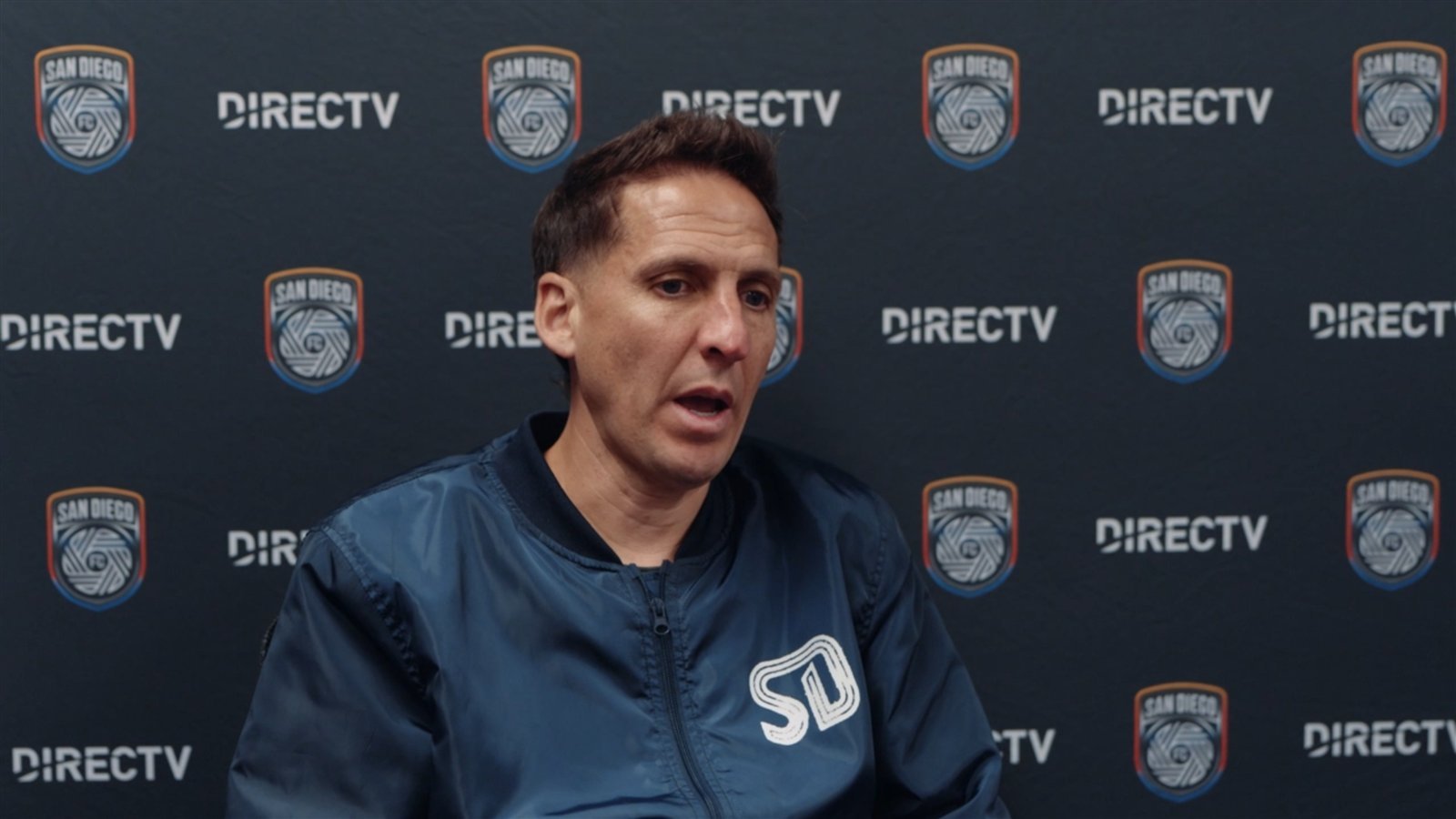 Mikey Varas, entrenador de San Diego FC, habla en conferencia de prensa frente a un fondo con el escudo del club.