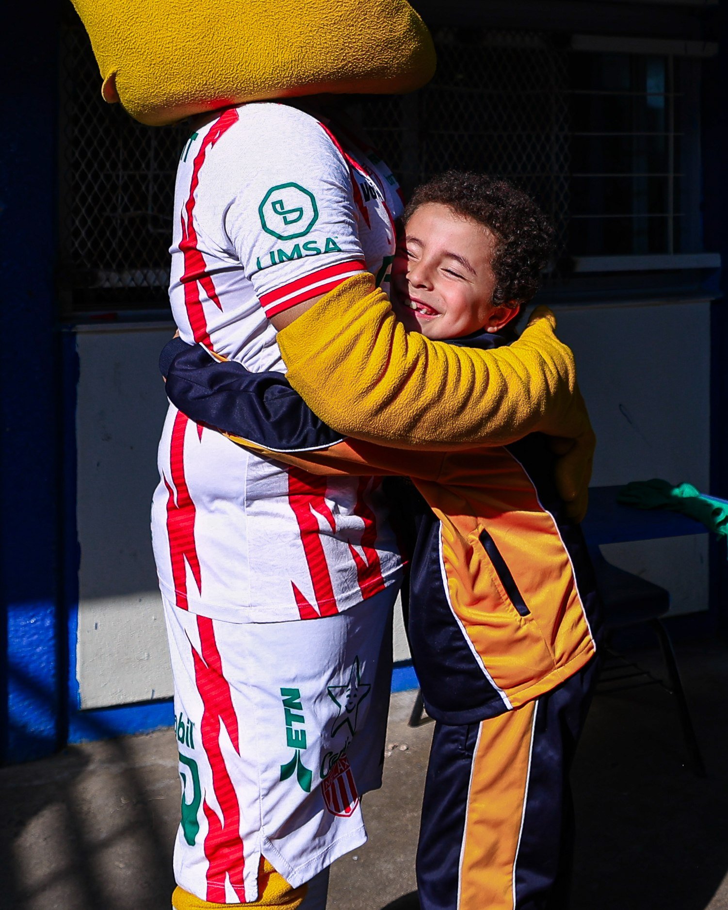 Un niño con uniforme escolar abraza sonriendo a la mascota del Club Necaxa, con la playera blanca y roja del equipo.