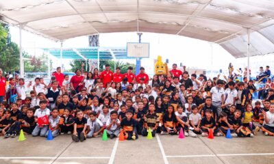 Jugadores de la Sub 17 del Necaxa, con playera roja, posan junto a la mascota del club y decenas de alumnos de la Escuela Primaria Alejandro Topete del Valle en el patio del plantel.
