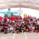 Jugadores de la Sub 17 del Necaxa, con playera roja, posan junto a la mascota del club y decenas de alumnos de la Escuela Primaria Alejandro Topete del Valle en el patio del plantel.