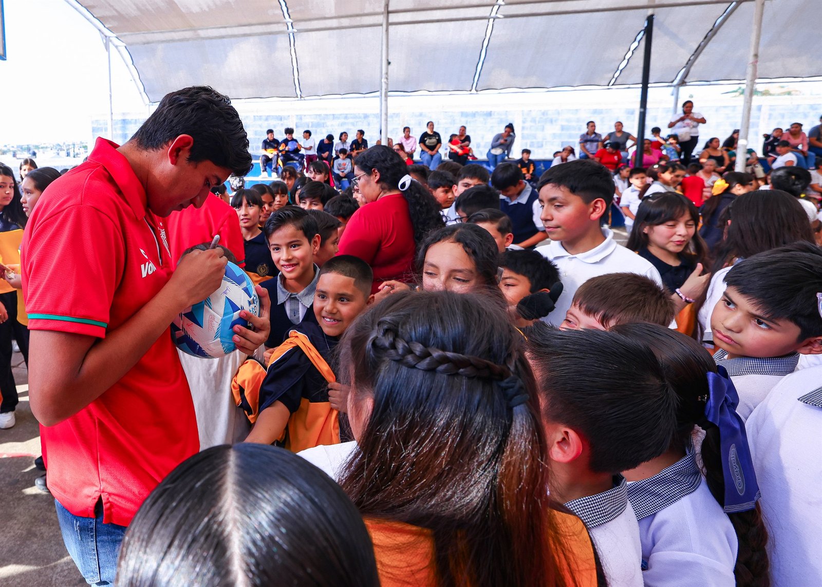 Un jugador de la Sub 17 del Necaxa con playera roja firma un balón rodeado de niñas y niños con uniforme escolar.