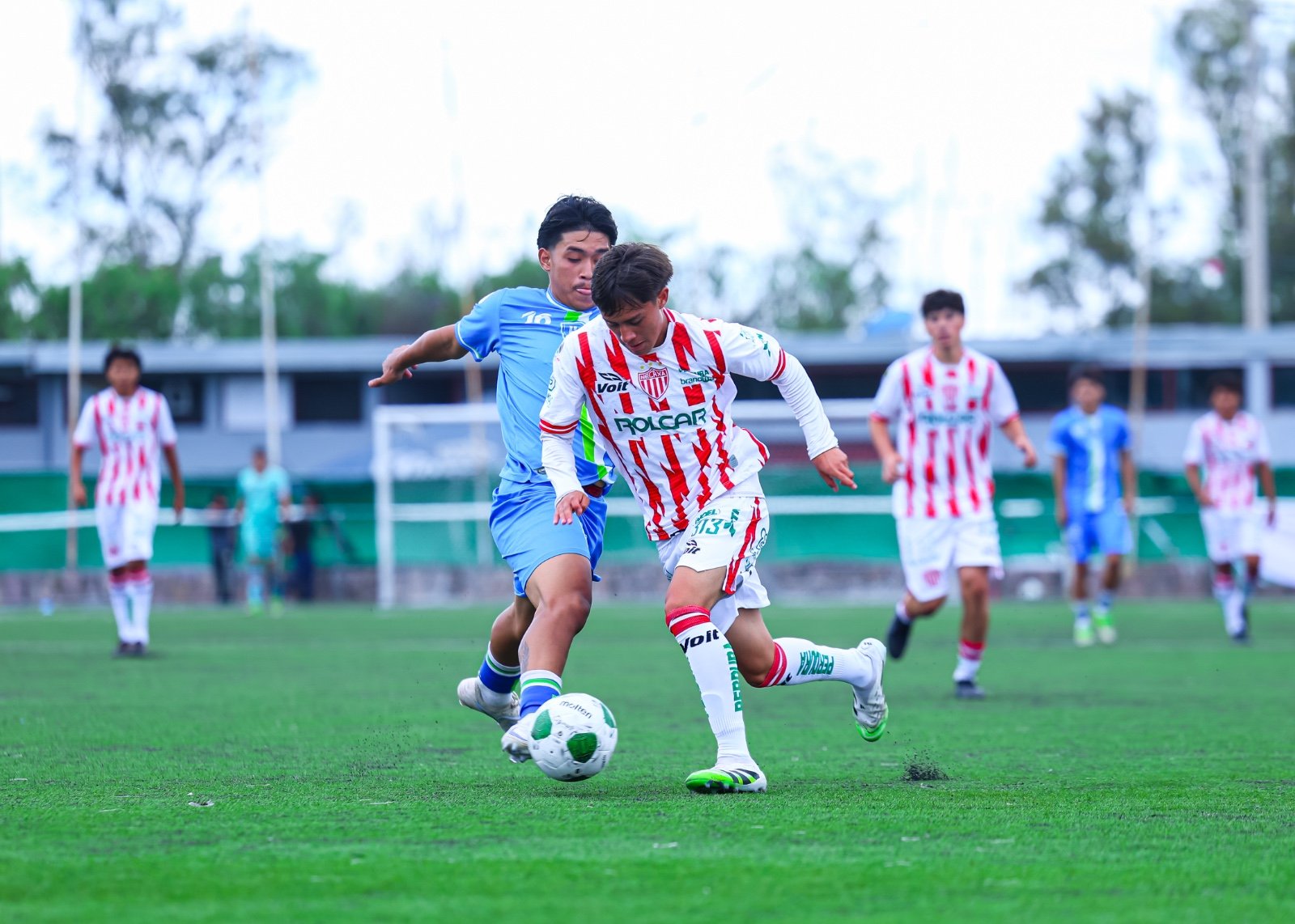 Un jugador de Necaxa con playera blanca y roja conduce el balón ante la marca de un rival con uniforme azul en un partido de la TDP en Casa Club Necaxa