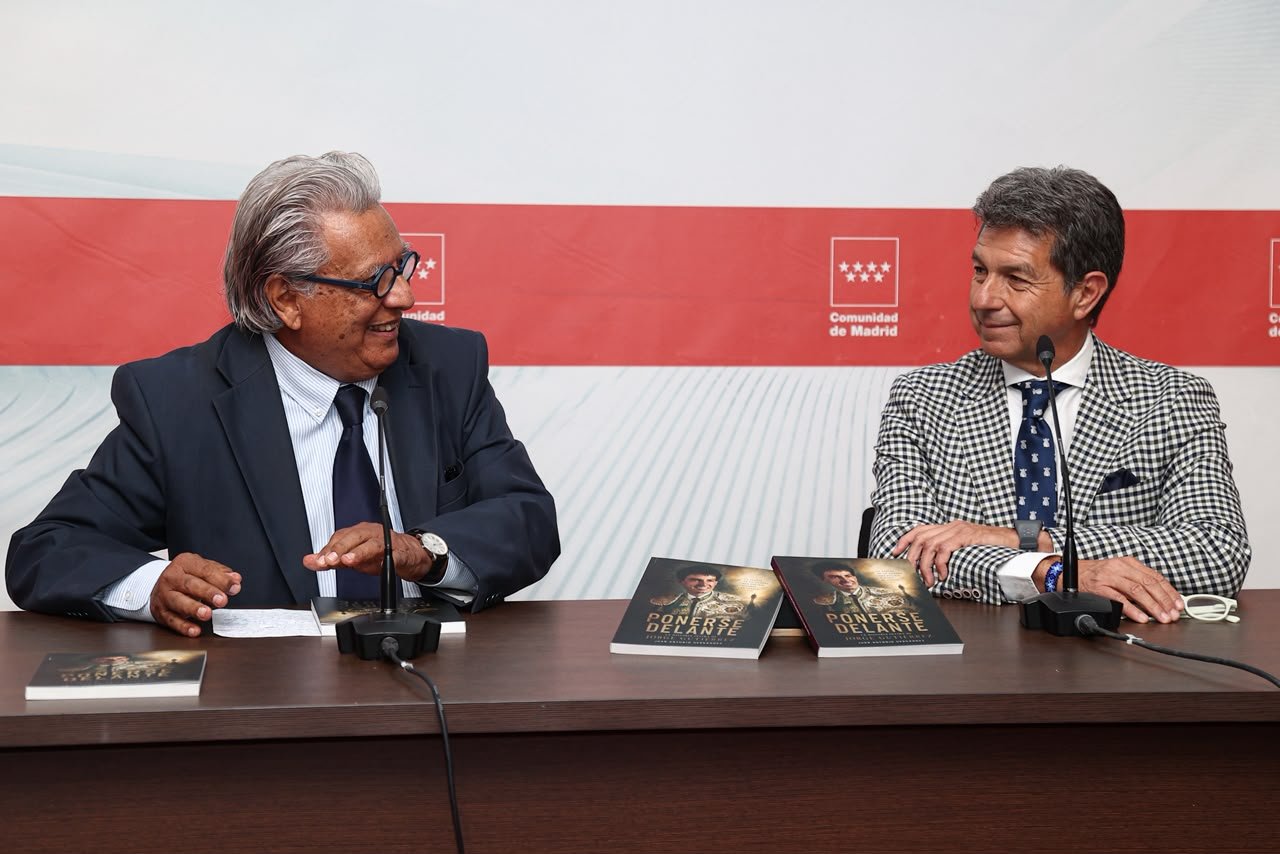 Dos hombres sentados en la mesa de la presentación del libro «Ponerse Delante», con ejemplares del libro sobre la mesa y el logotipo de la Comunidad de Madrid al fondo.